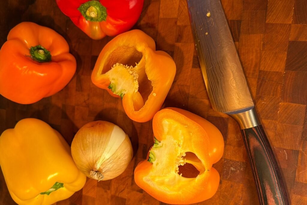 Bell peppers halved and seeded on a wooden cutting board next to a chef’s knife and whole onion.